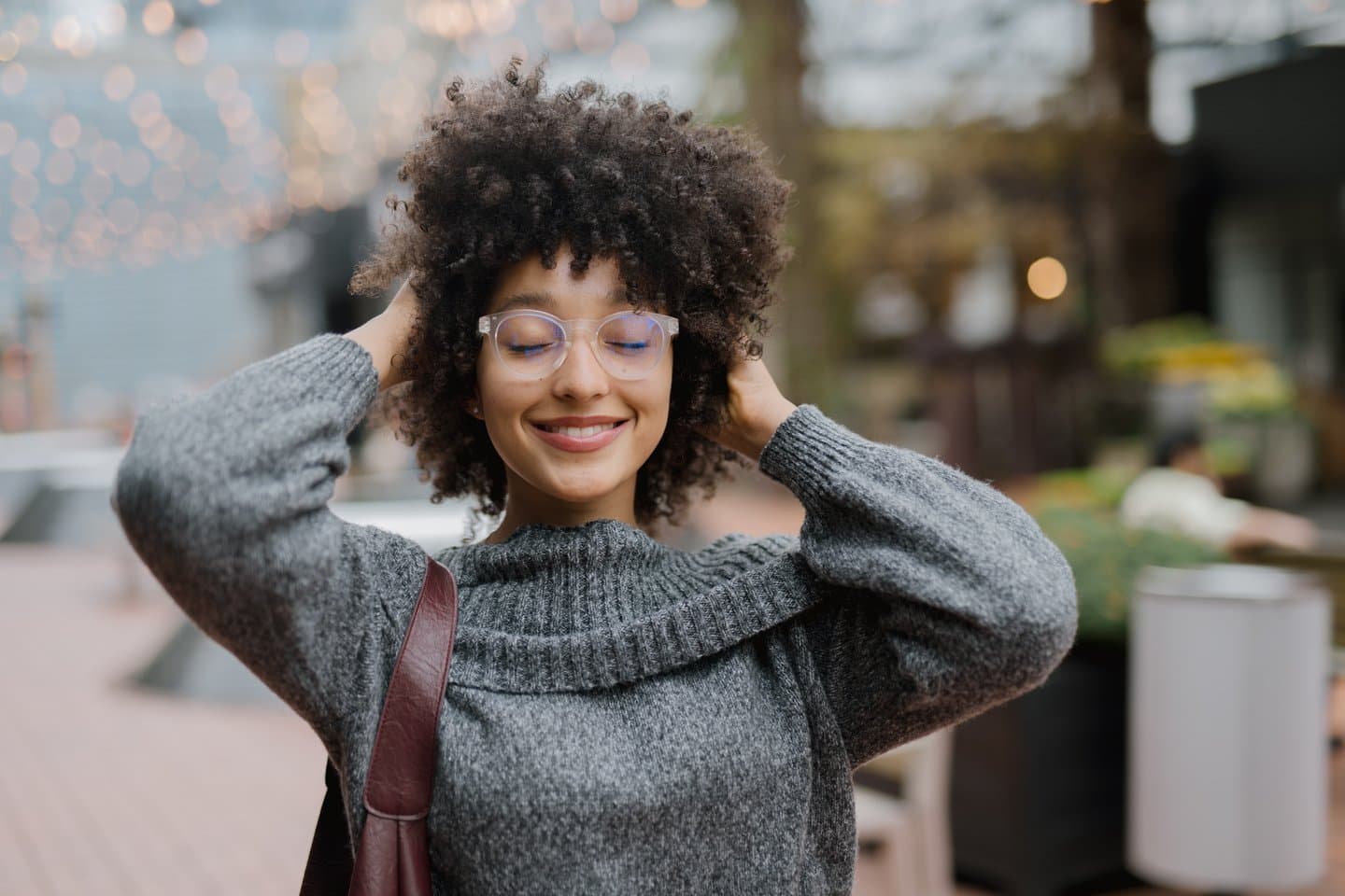 Smiling woman wearing bifocal lenses, grey sweater, and shoulder bag, standing outdoors with hands in curly hair.
